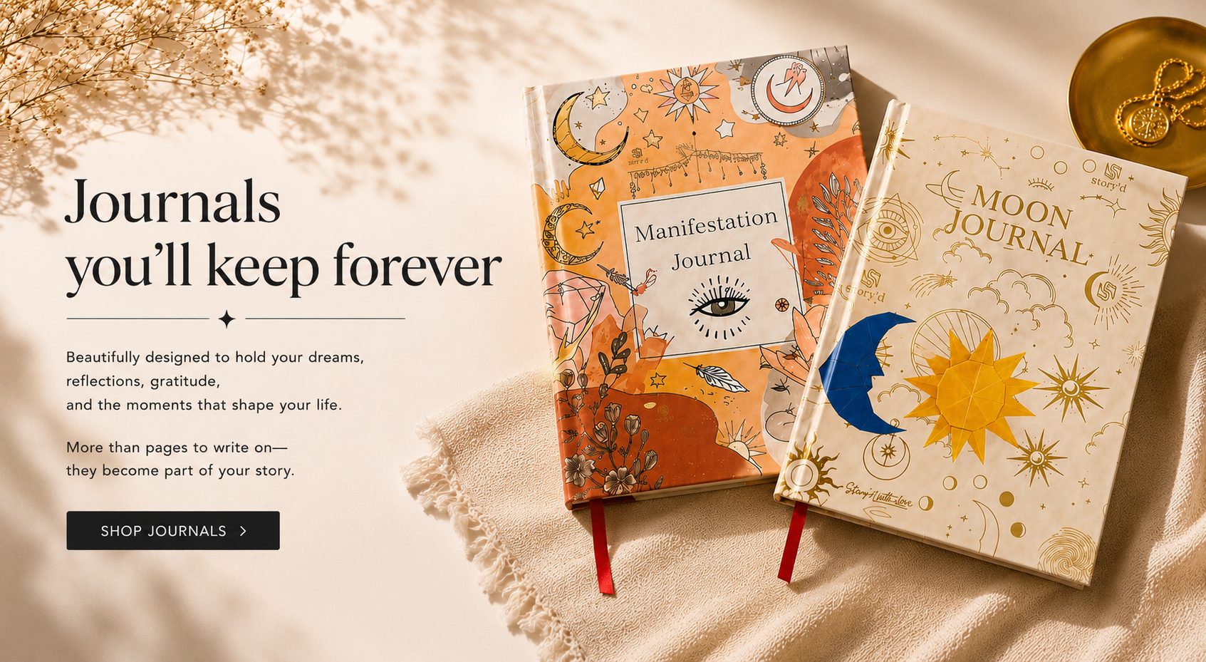 Two decorative journals displayed on a soft beige fabric background with a warm decorative scene. The left journal has a warm orange cover with celestial illustrations and the title 'Manifestation Journal.